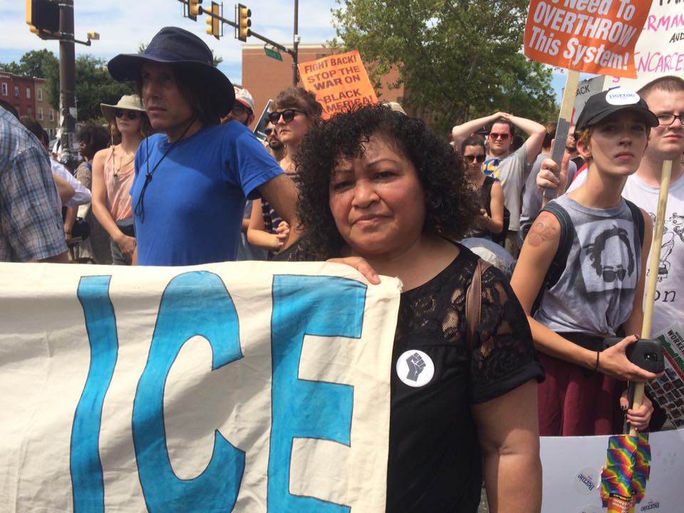 Portrait of a Latina woman holding the corner of a protest banner. ICE can be read in the corner of the banner.