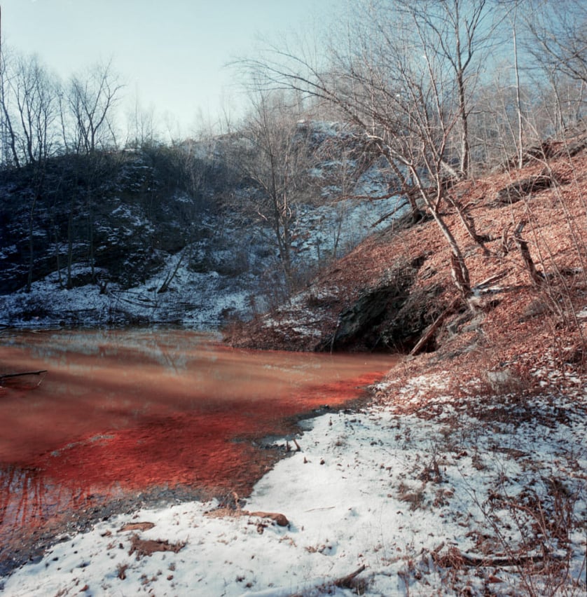 A photograph by Jeremy Blakeslee showing a reddish brown creek filled with coal mine tailings in the snowy woods of Centralia, PA.