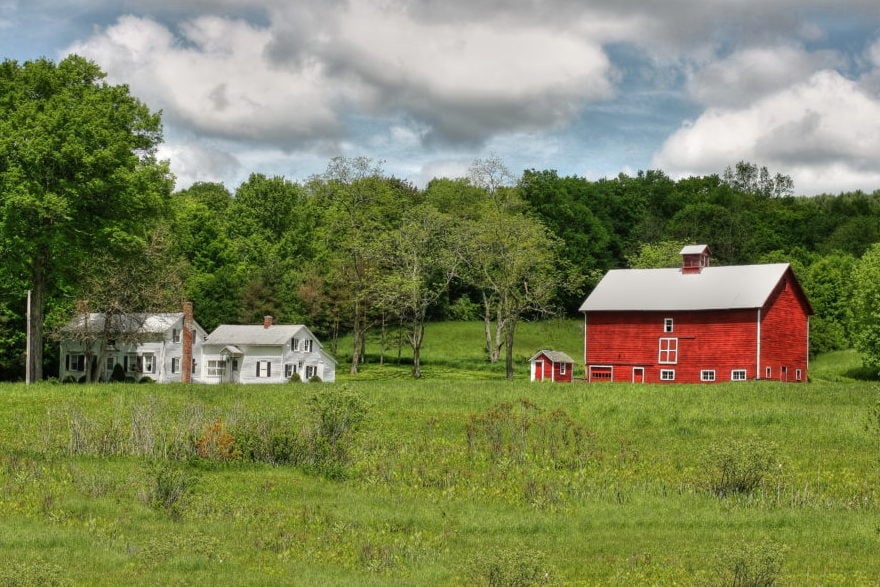 An upstate New York farm featuring a white house and red barn in front of a line of trees.