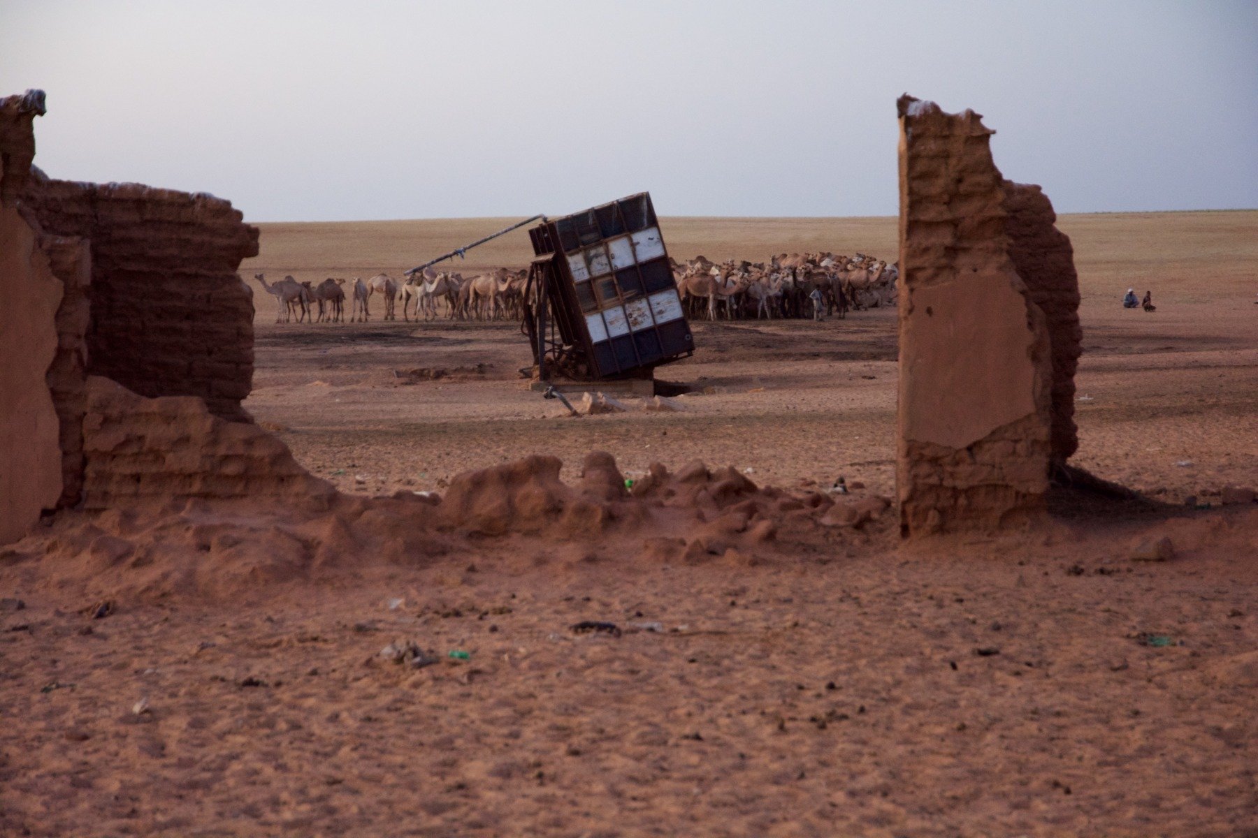 A destroyed water station in the desert. In the far background, a herd of camels.