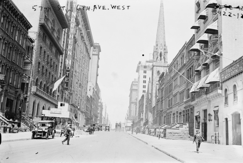 A black and white photo of a New York street. A church steeple pokes out from behind walk-ups and tenements.