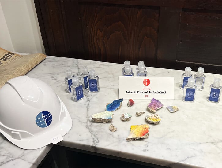 On a white marble counter in the museum's gift shop: a hard had with the logo of the Victims of Communism Foundation; mini bottles of hand sanitizer; pieces of the Berlin wall. 