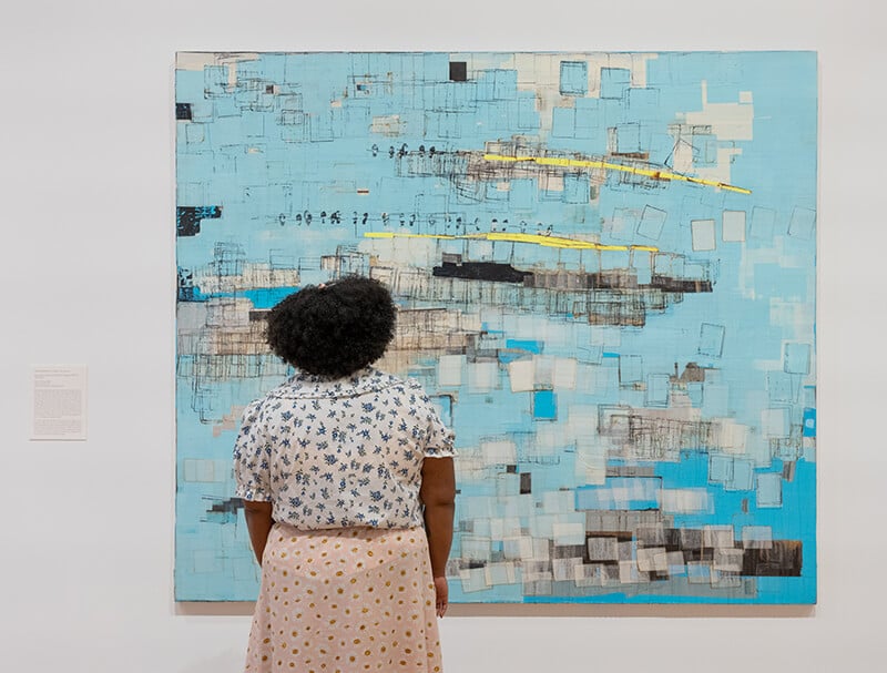 A woman gazes at a painting on a wall: abstract, comprised largely of teal blue squares, with gold and black accents.