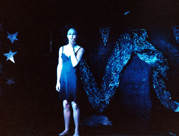 A bald transgender woman stands on a stage in a black slip smoking a cigarette.