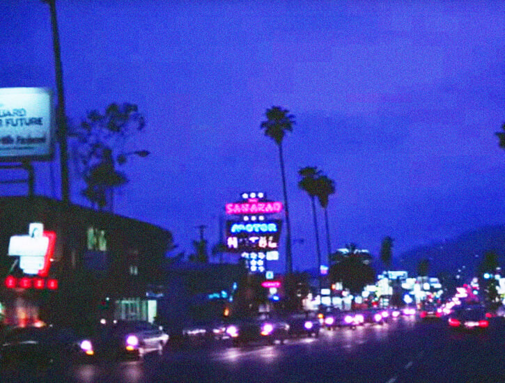 A line of cars in traffic at night, with palm trees and neon signs in the background.