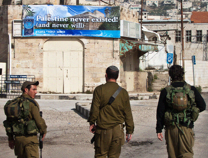 Three soldiers stand before a sign that says "Palestine never existed! (and never will)"