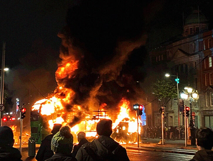 A bus in flames on a city street with a crowd of onlookers in the foreground.