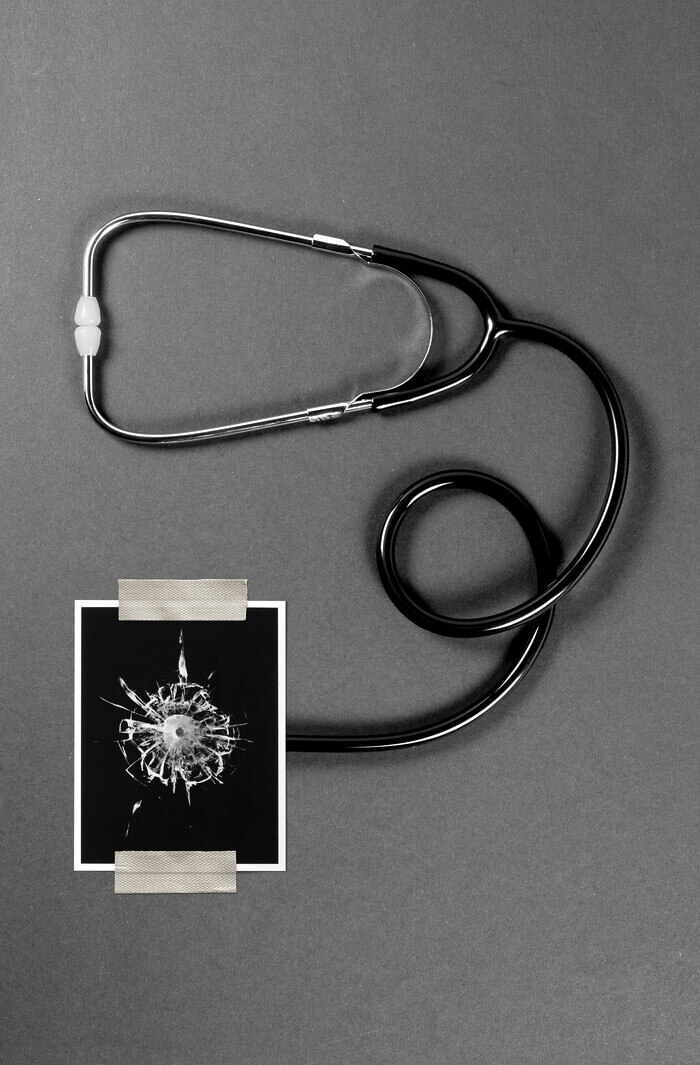 A stethoscope lies curled on a table, and the bell is covered by a photo of broken glass that appears to have had a bullet go through it.