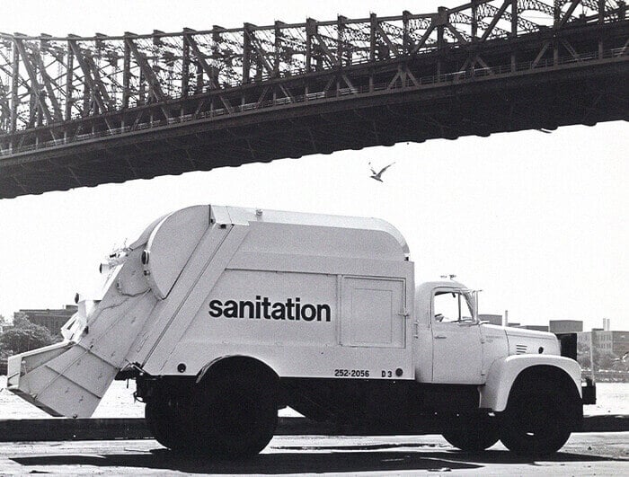 A white garbage truck with the word "sanitation" is parked before a bridge in New York City.