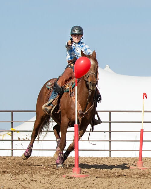 A red balloon on a stick stands dead center, and directly behind it is a grade school child wearing a helmet, riding a horse, and pointing a gun at the balloon.