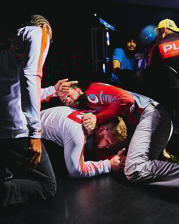 A photo of two men wrestling on a black floor, with a third man kneeling over them and pointing.