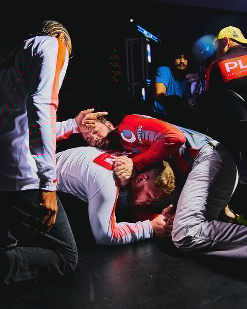 A photo of two men wrestling on a black floor, with a third man kneeling over them and pointing.