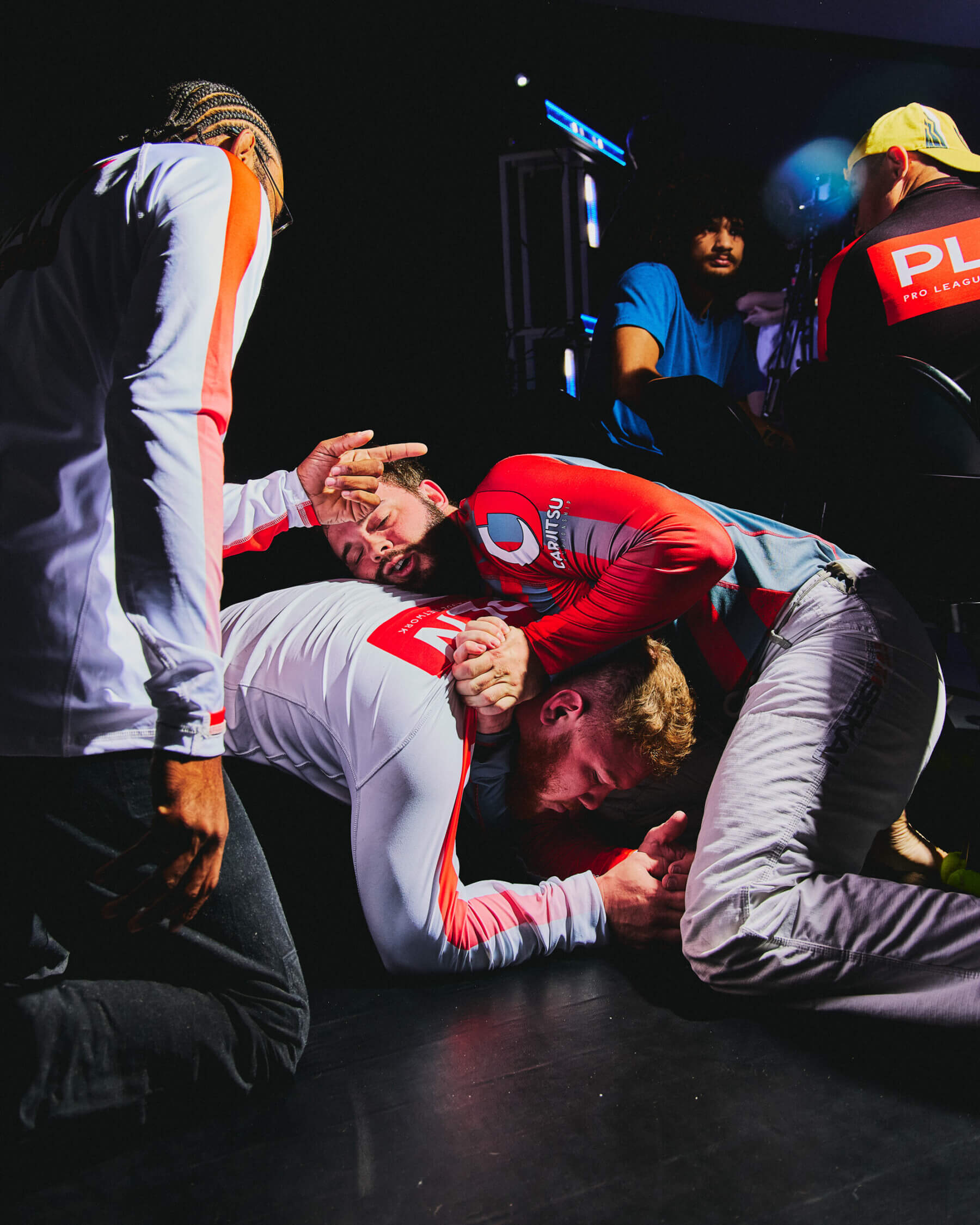 A photo of two men wrestling on a black floor, with a third man kneeling over them and pointing.