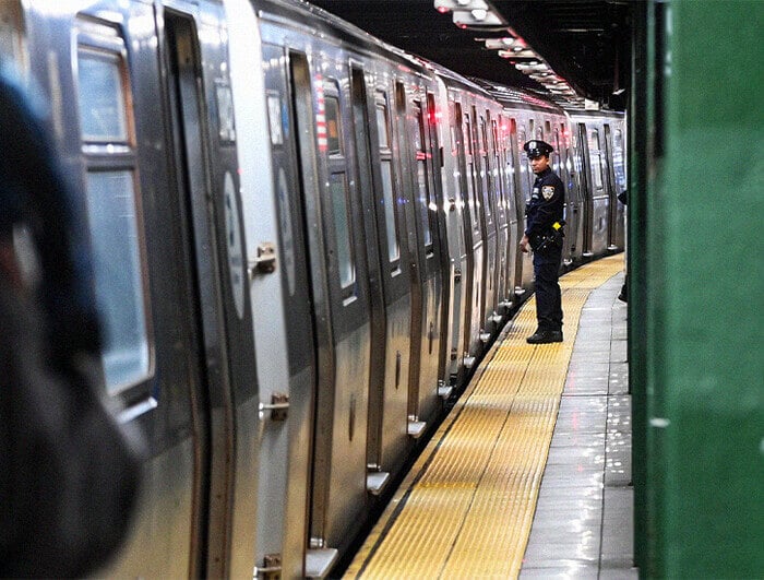 An NYPD officer stands on a subway platform while a subway comes into the station.