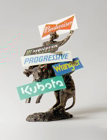 A staged photograph of a bronze statue of a cowboy riding a bull standing on its back legs is placed before a white seamless backdrop. There are signs for Budweiser, Monster Energy, Progressive Insurance, Wrangler, and Kubota resting on it.