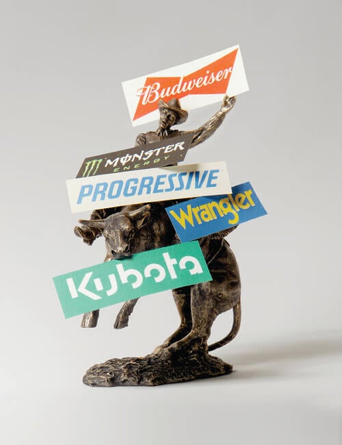 A staged photograph of a bronze statue of a cowboy riding a bull standing on its back legs is placed before a white seamless backdrop. There are signs for Budweiser, Monster Energy, Progressive Insurance, Wrangler, and Kubota resting on it.