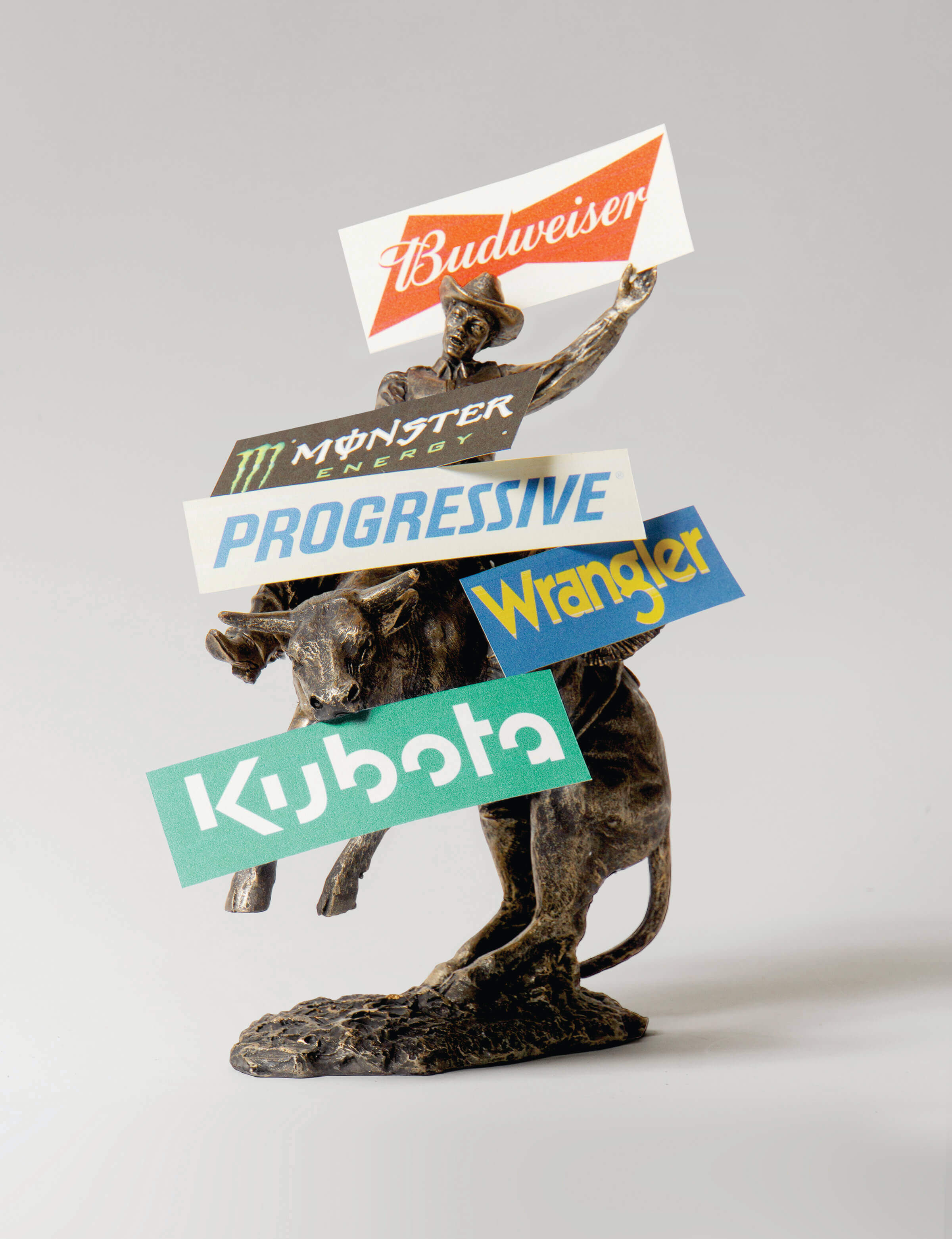 A staged photograph of a bronze statue of a cowboy riding a bull standing on its back legs is placed before a white seamless backdrop. There are signs for Budweiser, Monster Energy, Progressive Insurance, Wrangler, and Kubota resting on it.
