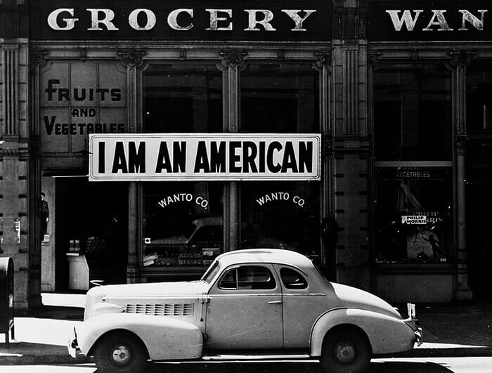 A large sign reading "I am an American" placed in the window of a store in. A parked car is in the foreground.