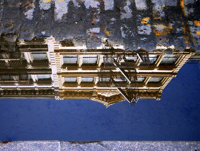 A building's facade in New York is reflected in a puddle of water on the street below.