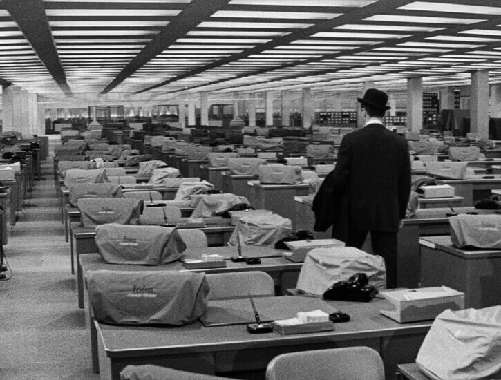 A man in a black suit and hat walks away from the camera, down a row of empty desks in an office.