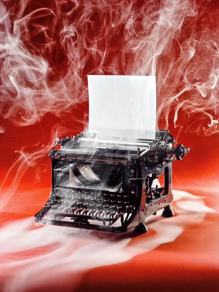 A photograph of an old-fashioned black typewriter sits on a red backdrop with smoke all around it. There’s a blank piece of paper rolled into the typewriter.