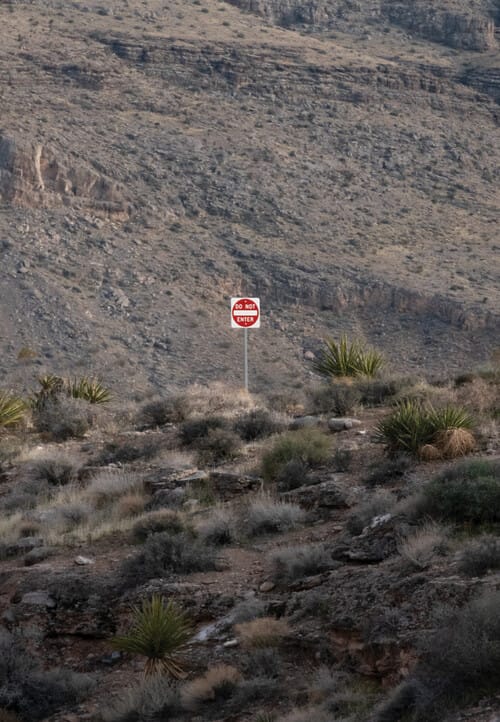 A photograph of a zoomed out sign that reads “DO NOT ENTER” standing in the middle of large hills with small brown and dark green shrubbery around it.