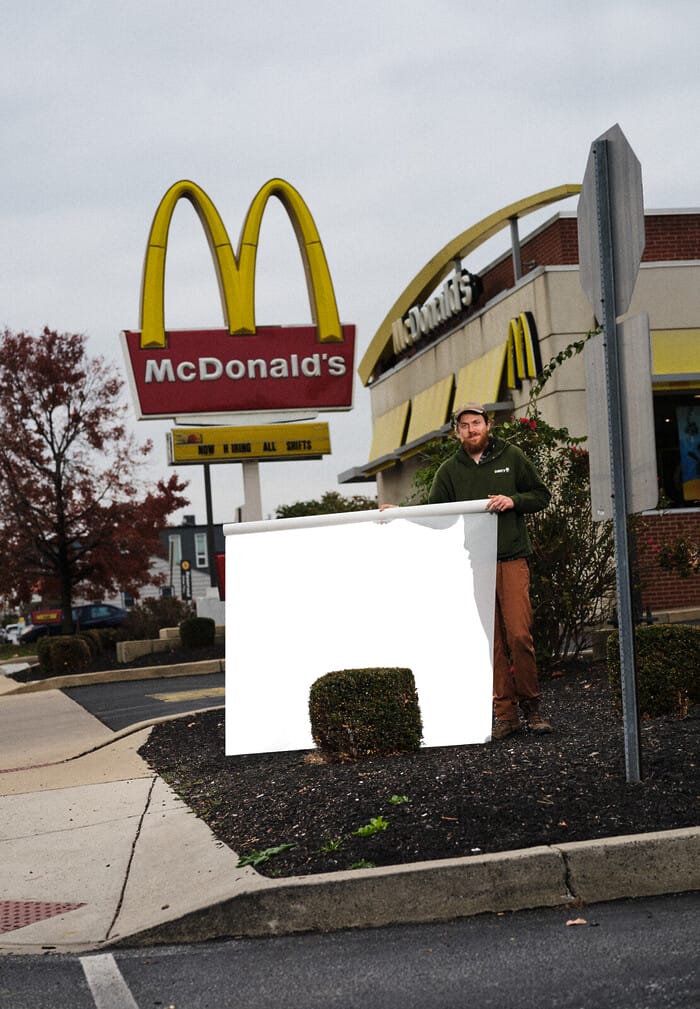 A photograph of a man wearing a baseball cap, green hoodie, tan pants, and boots holds a white scroll in front of a short manicured bush in a parking lot. Behind him is a large McDonald’s sign on the left, and a McDonald’s building on the right.