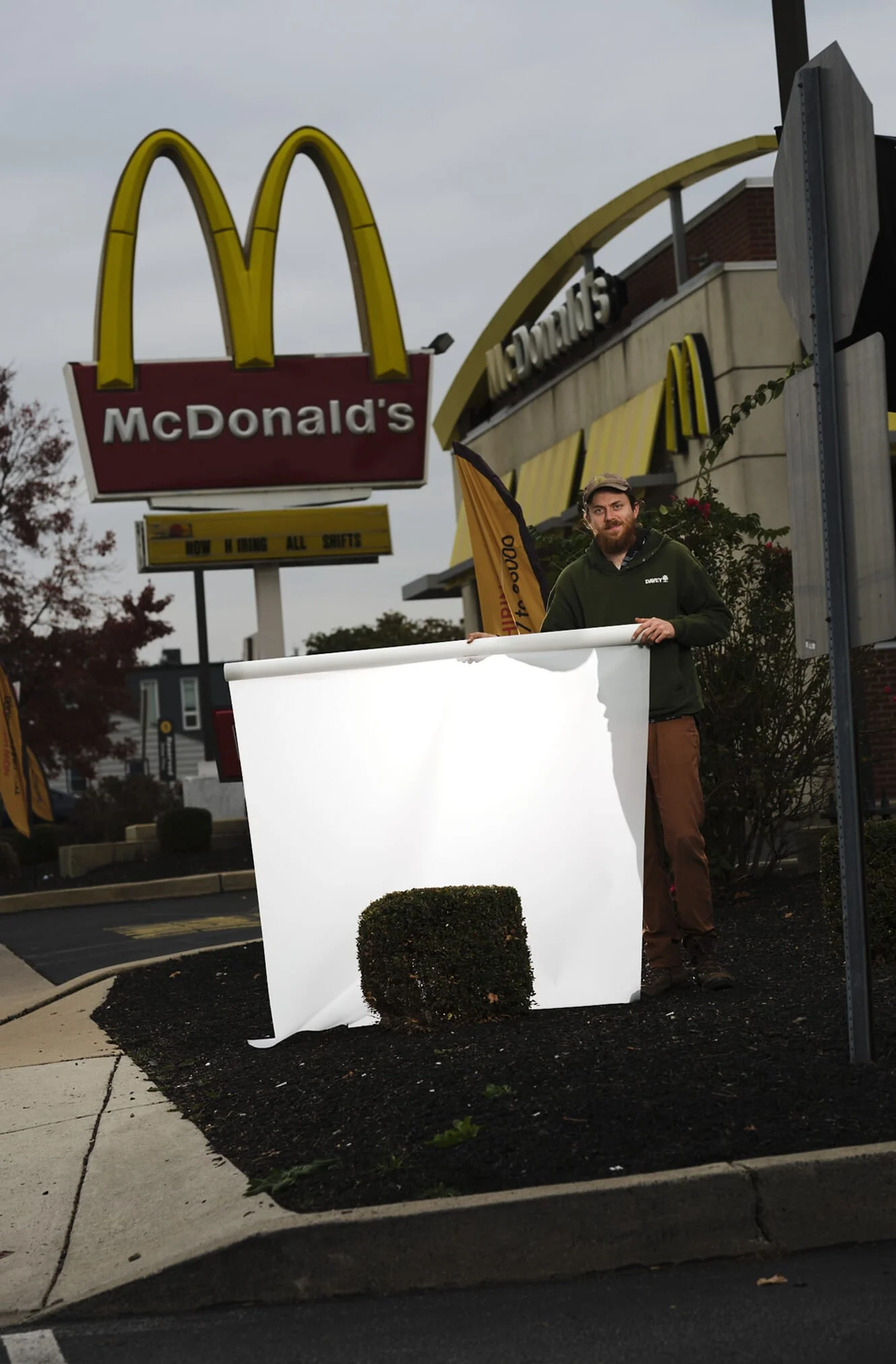 A photograph of a man wearing a baseball cap, green hoodie, tan pants, and boots holds a white scroll in front of a short manicured bush in a parking lot. Behind him is a large McDonald’s sign on the left, and a McDonald’s building on the right.