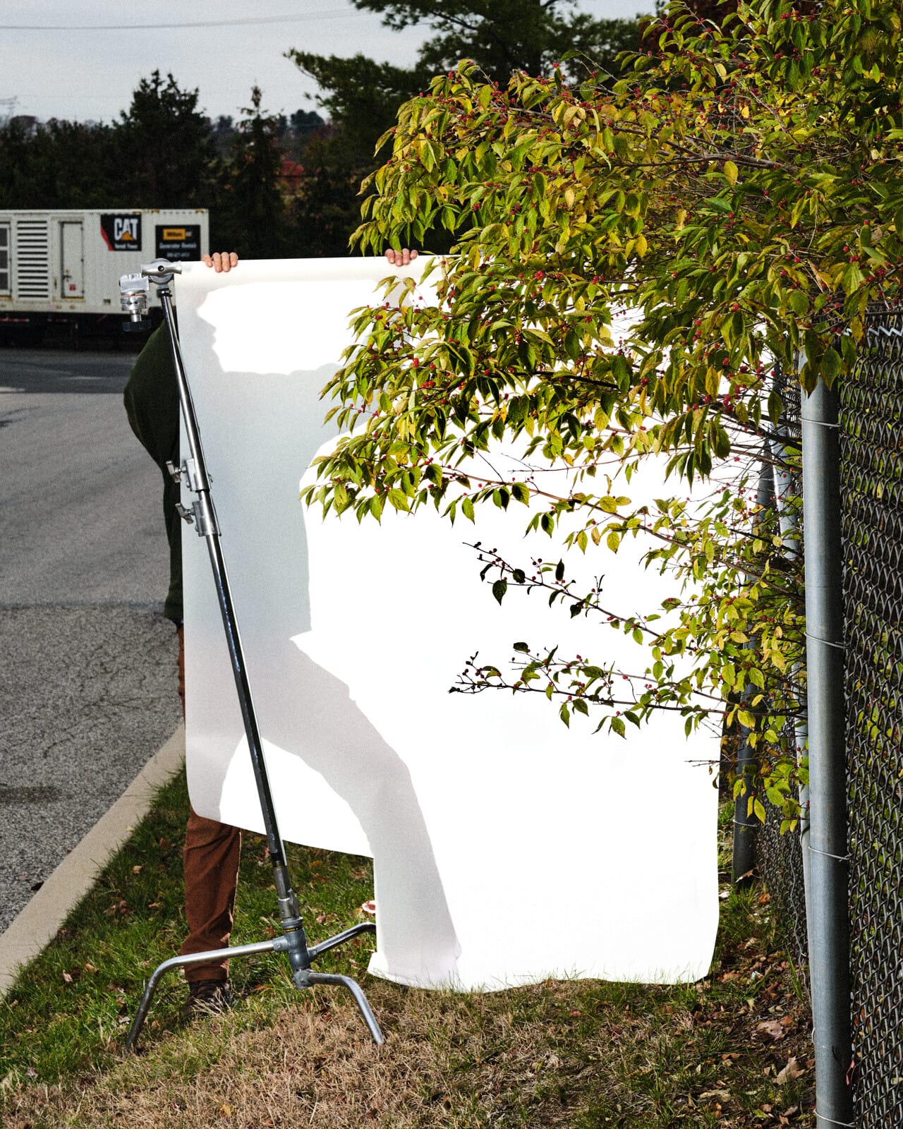 A white screen is held by a man, along with a tall metal apparatus, by the side of a road. Before the screen, a tree grows through a chain-link fence.