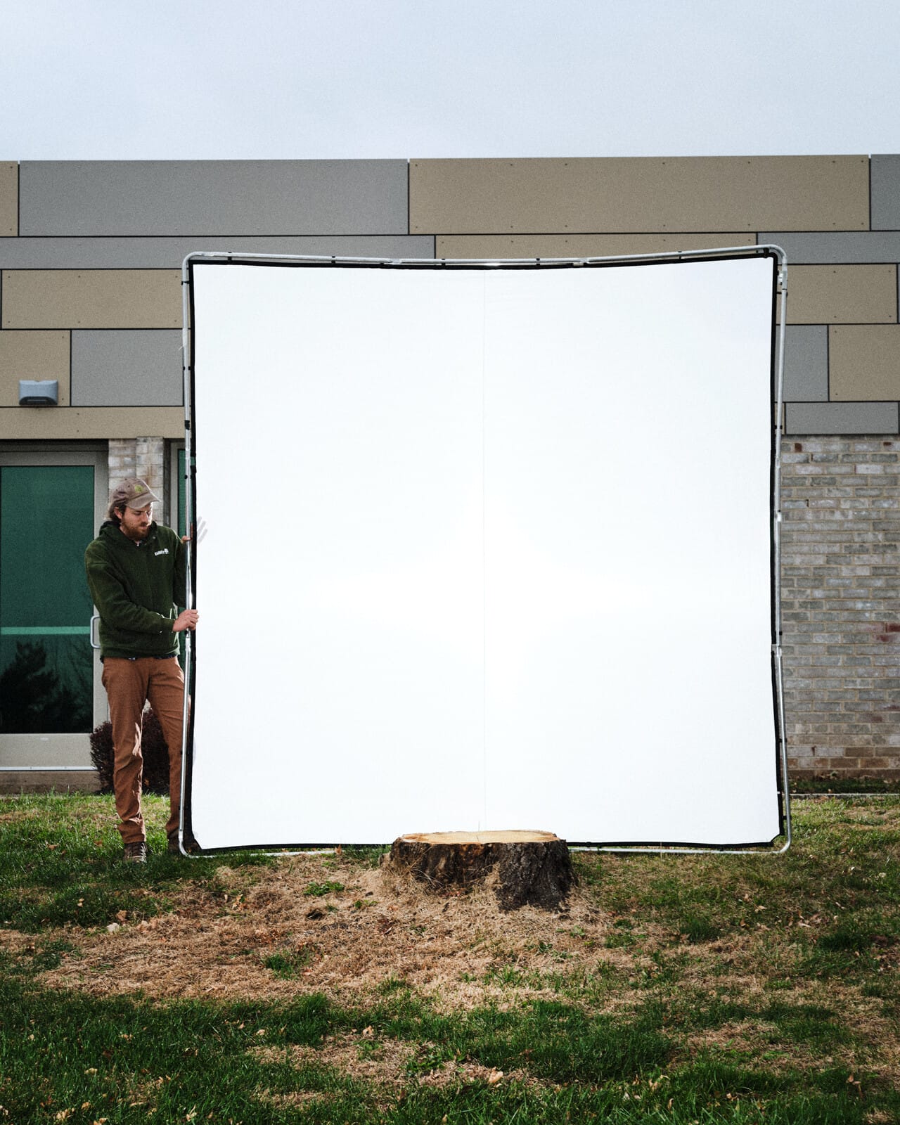 A photograph of a man holding the left side of a large square screen. In front of the screen is a freshly cut tree stump. Behind the screen is a muted gray-and-beige concrete-and-brick building.