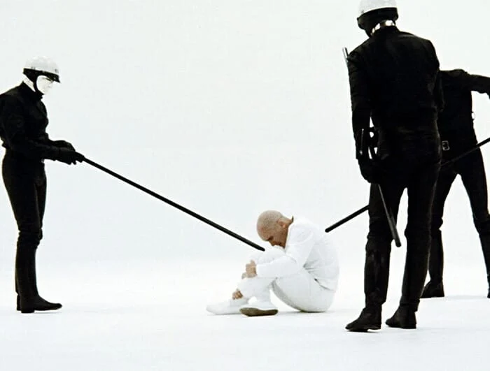 In an all white room, three police officers dressed in all black surround a man sitting on the ground, who wears all white.