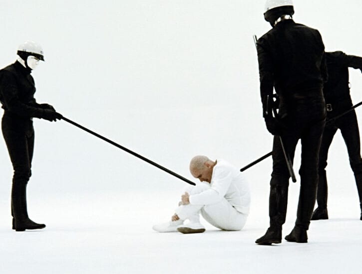 In an all white room, three police officers dressed in all black surround a man sitting on the ground, who wears all white.