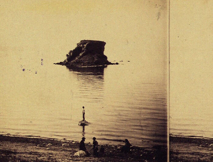 In the foreground, a man stands on a rock. In the distance, the Black Rock of the Great Salt Lake emerges from the water.