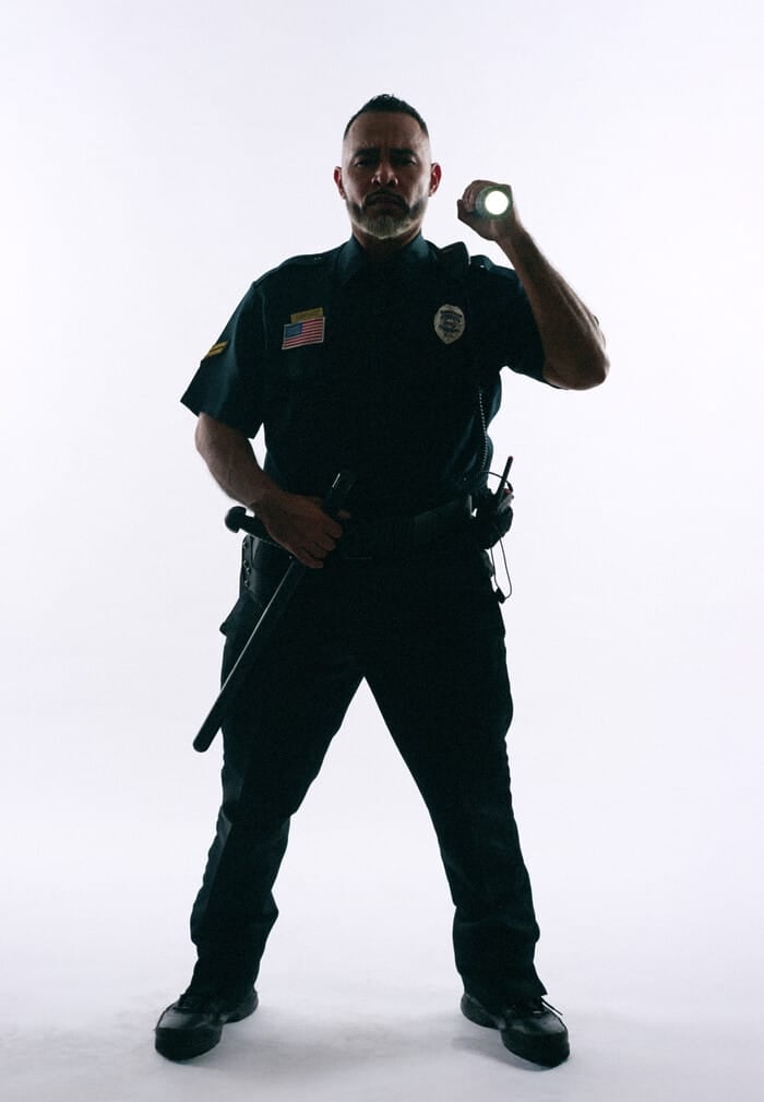A uniformed police officer stands with one hand on his baton and the other hand pointing a flashlight as though he’s investigating something.