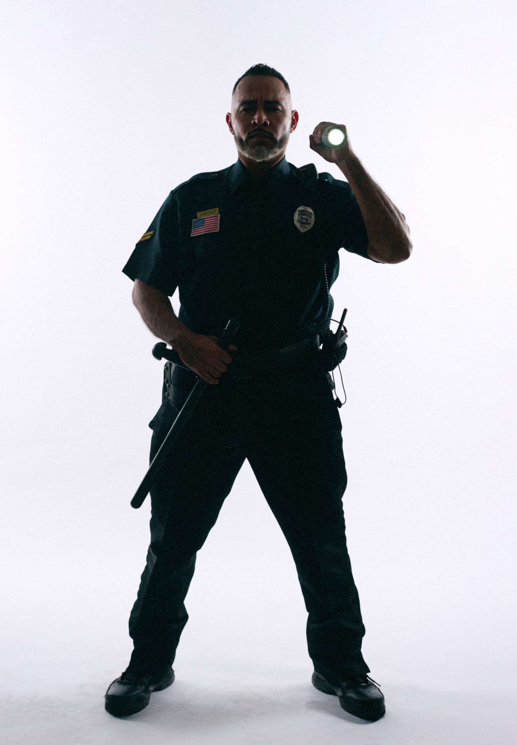A uniformed police officer stands with one hand on his baton and the other hand pointing a flashlight as though he’s investigating something.