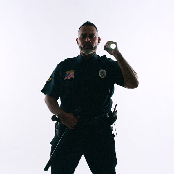 A uniformed police officer stands with one hand on his baton and the other hand pointing a flashlight as though he’s investigating something.