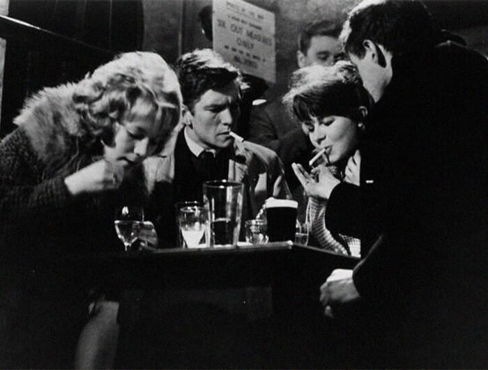 At a table in a bar, four people sit: from left, a woman, a man and woman, and another man. Two are smoking.