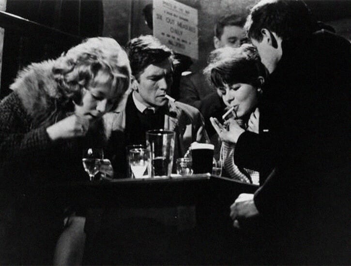 At a table in a bar, four people sit: from left, a woman, a man and woman, and another man. Two are smoking.