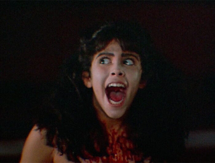 A preteen child with long brown hair screams. There is blood on their decolletage.