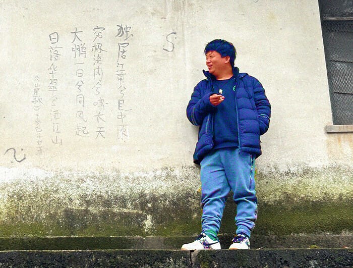A man in a blue jacket stands before a wall on which a limerick has been written in Chinese characters.