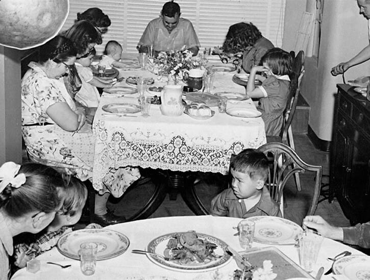 A dining room has two tables: in the back, eight people are seated, heads bowed in prayer. In the foreground, four children are seated.