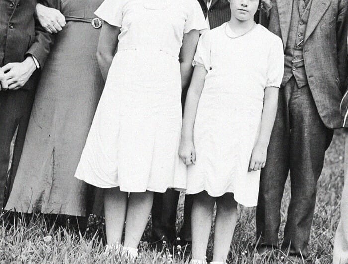 A black and white portrait of a family—with two young girls dressed in white at center. The faces of all are obscured.