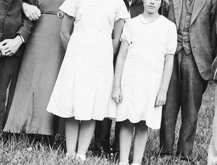 A black and white portrait of a family—with two young girls dressed in white at center. The faces of all are obscured.