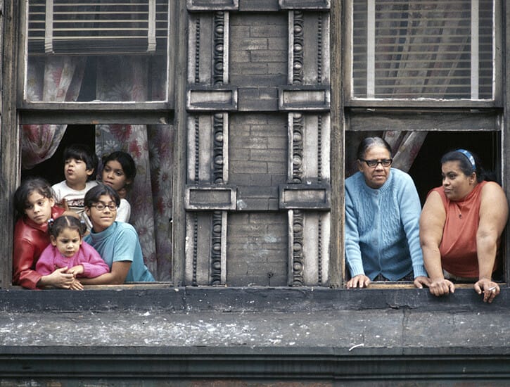 A photo of two windows of an apartment building: in the left, a group of five children look out; in the right, two older women looking out.