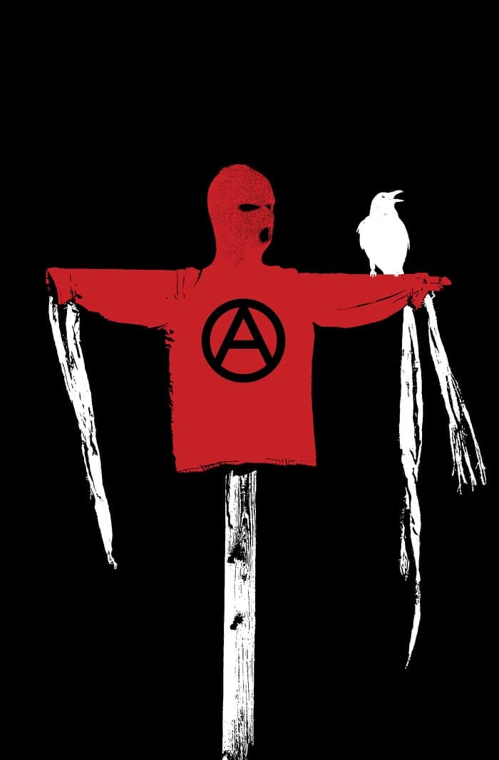 A scarecrow wearing a red hoodie with an anarchy symbol on the chest stands on a pole against a black background. A white bird sits on one of its arms.